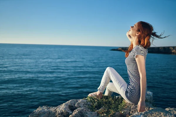 Woman in a t-shirt and trousers sits on a stone near the sea in summer — Stock Photo, Image