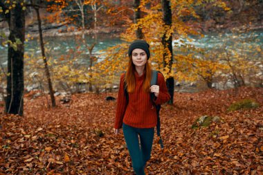 Kırmızı kazaklı ve şapkalı bir gezgin, sonbahar ormanında dağın tepesindeki manzaranın yanında yürüyor.