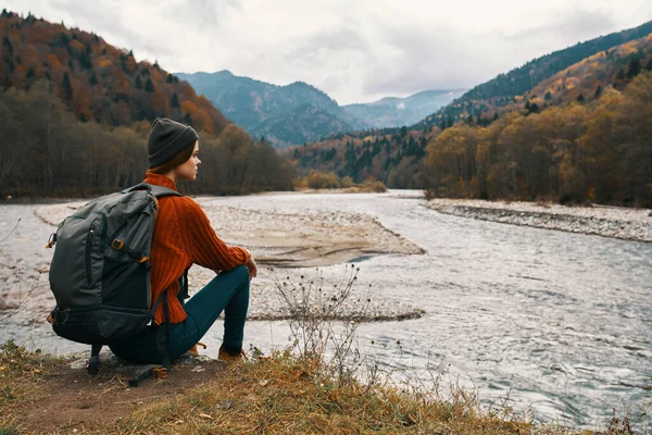 Femme avec un sac à dos dans une veste et un chapeau sur la rive de la rivière dans la vue sur les montagnes — Photo