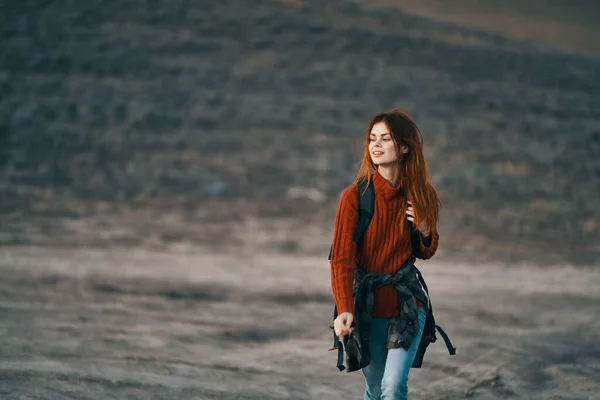 Cestovatelka v červeném svetru s batohem na zádech a v džínách kráčí v přírodě v horách — Stock fotografie
