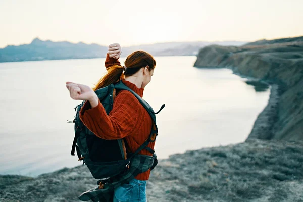 Felice donna in un maglione rosso con uno zaino sulla schiena che riposa nella natura vicino al mare e alle montagne in lontananza — Foto Stock