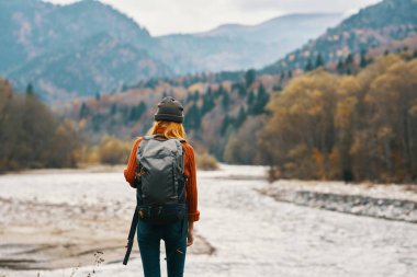 Nehir kıyısında sırt çantalı, uzaktaki dağlarda kırmızı kazaklı bir kadın.