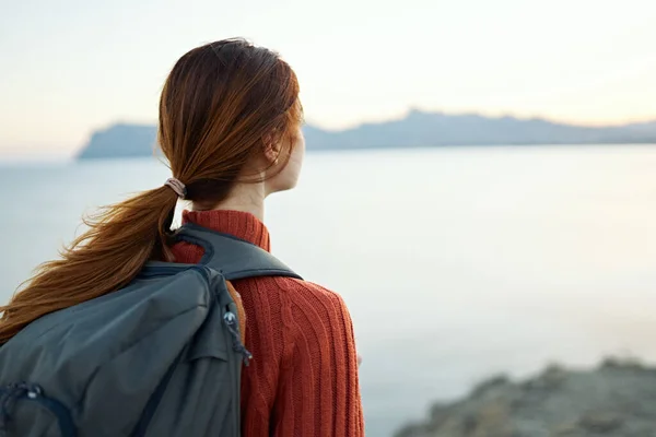 Donna turista in un maglione arancione e con uno zaino sulla schiena sta riposando vicino al mare e le montagne in lontananza — Foto Stock