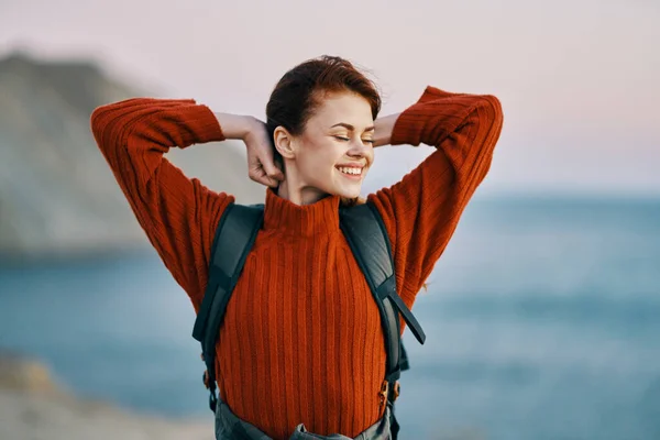 Vrouw in een rode trui met een rugzak op haar rug rusten in de bergen buiten in de buurt van de oceaan — Stockfoto