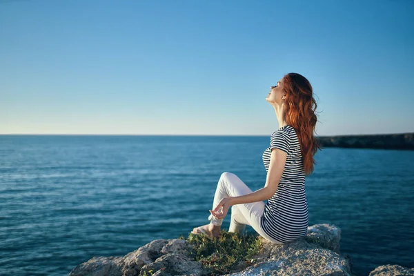 Szczęśliwa kobieta w t-shirt spodnie siedzi na dużym kamieniu w pobliżu morza w lecie — Zdjęcie stockowe
