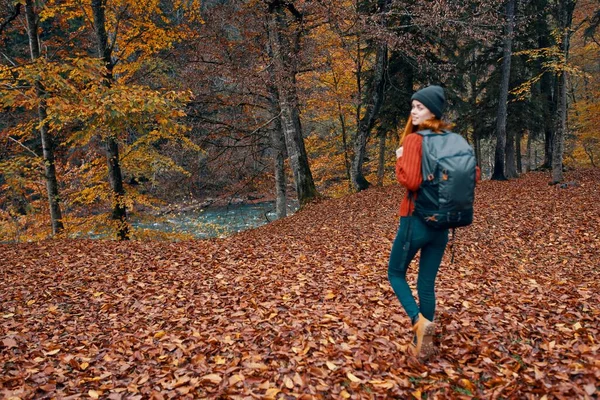 Heureuse jeune femme avec un sac à dos en bottes de jeans et un pull marchent dans la forêt d'automne près des grands arbres — Photo