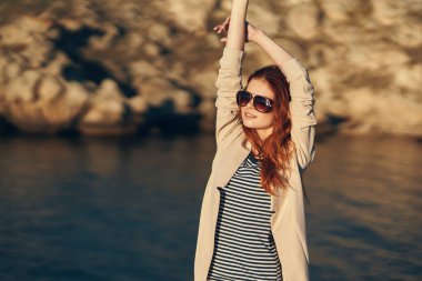 Tişörtlü kadın ellerini başının üstünde tutuyor. Dağlarda, denizin kenarında.