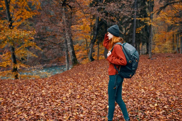 Vrouw met een hoed in een rode trui en jeans wandelingen in het park met een rugzak op haar rug reizen toerisme herfst landschap — Stockfoto
