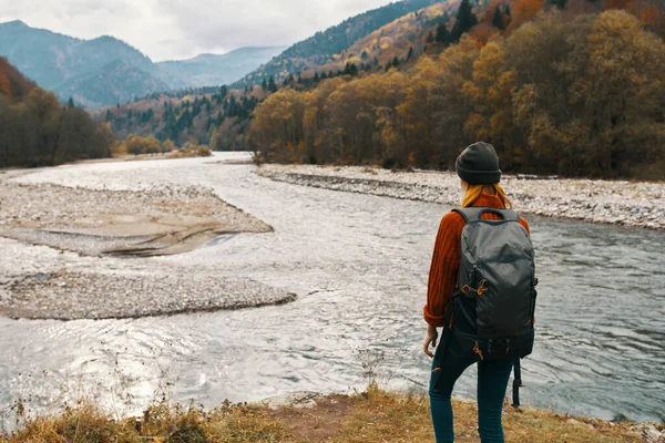 Uma mulher com uma jaqueta vermelha e uma mochila nas costas estão andando nas margens do rio nas montanhas — Fotografia de Stock