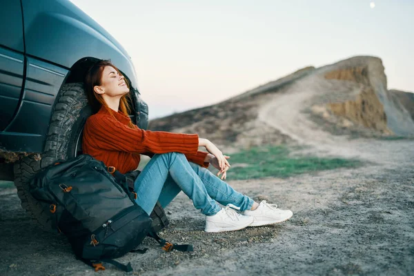 the traveler sits near the car in nature in the mountains and a backpack on the side