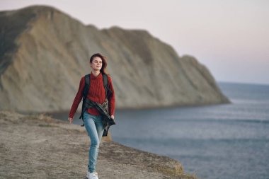 Sırtında sırt çantası olan bir kadın dağlarda deniz kenarında doğa yürüyüşü yapıyor.