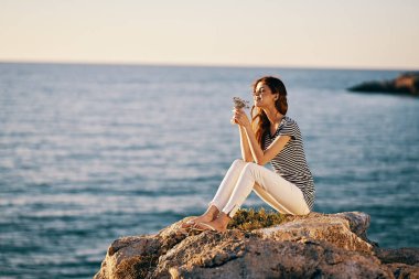 Bir kadın deniz kenarındaki bir kayanın üzerinde pantolon ve tişörtle oturuyor.