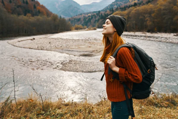 Mujer en un suéter rojo y gorra con una mochila en la espalda en las montañas cerca del río en la naturaleza —  Fotos de Stock