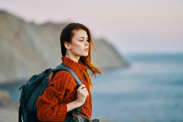 Viaggiatore in un maglione rosso vicino al mare in natura con uno zaino sulla schiena e montagne sullo sfondo — Foto Stock