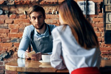 Kafe masasında bir fincan kahve ve önlüklü bir adam ve kırmızı etekli bir kadın.