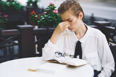 Restoranda bir masada kitap okuyan bir kadın eğitim duyguları arka planda çiçekler gözlüyor.