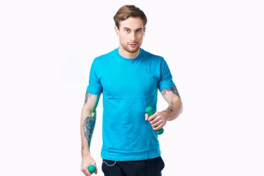 Elinde dambıllar olan mavi tişörtlü bir sporcu spor salonu dövmesi yaptırıyor.