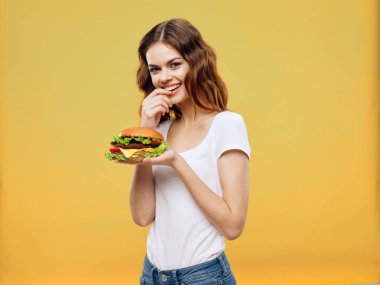 Beyaz tişörtlü hamburgerci kadın elinde fast food diyeti sarı arka plan