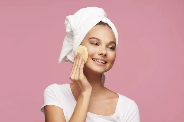 Happy woman with towel on head wipes face with soft sponge on pink background — Stock fotografie