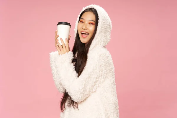 Feliz mujer asiática con una taza de café sobre un fondo rosa y un suéter con capucha caliente —  Fotos de Stock