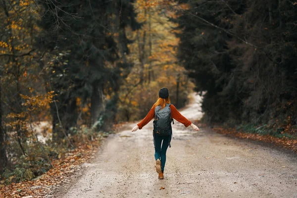 Mujer en chaqueta vaquera con una mochila camina por la carretera en el bosque de otoño — Foto de Stock