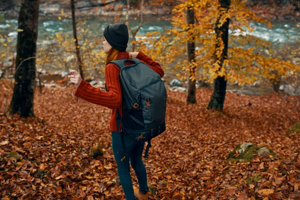 Vrouw in een trui met rugzak en in spijkerbroek wandelt door het herfstbos in de bergen bij de rivier — Stockfoto