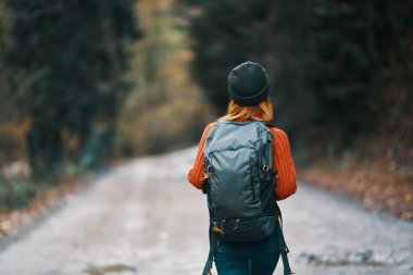 Sırt çantalı, şapkalı ve turuncu kazaklı bir kadın sonbahar ormanı manzaralı yolda.