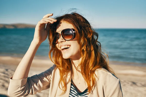 Kobieta w czerwonych włosach i okularach słonecznych śmieje się w przyrodzie na plaży w pobliżu morza — Zdjęcie stockowe