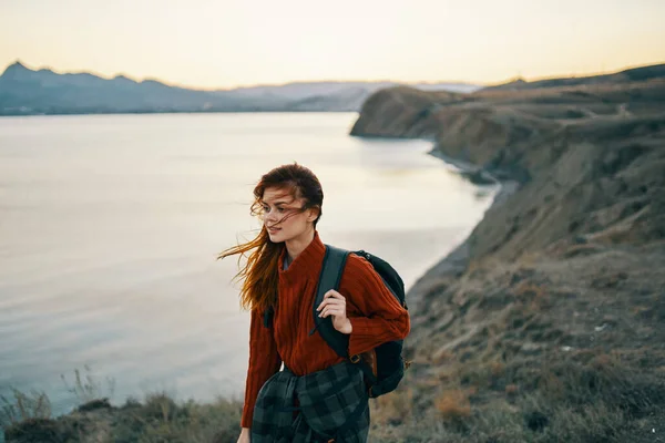 Krásná žena ve svetru s batohem v horách v blízkosti moře a vysoké skály západ slunce příroda — Stock fotografie
