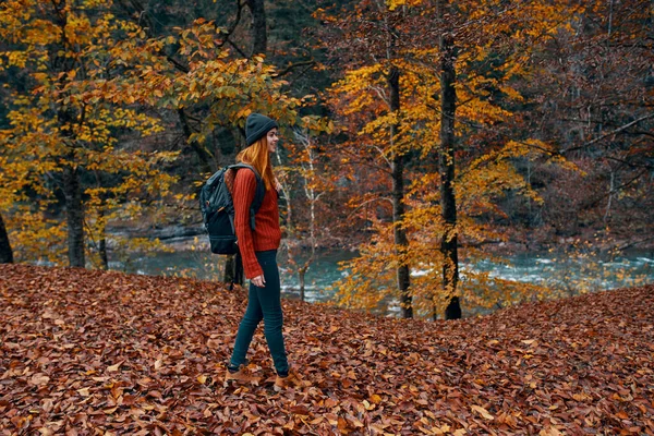 Vrouw in de herfst in het park met gevallen bladeren en een rugzak op haar rug rivier op de achtergrond — Stockfoto