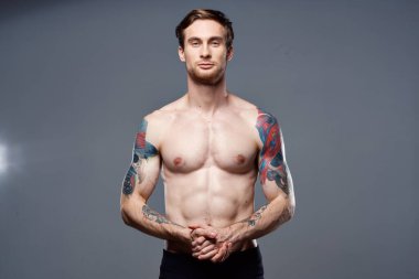 Kollarında çıplak şişirilmiş gövde dövmeleri olan yakışıklı bir adam ellerini kemerinde tutuyor.