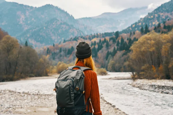 Mujer se para en la orilla del río y mira a las montañas en la distancia paisaje otoño mochila turismo modelo —  Fotos de Stock