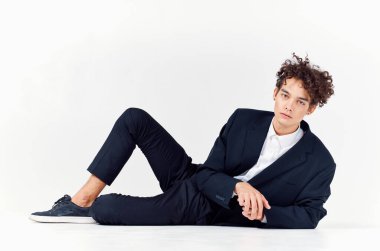 Kıvırcık saçlı bir adam yerde takım elbisesinin içinde uzanır. Parlak bir odada ve spor ayakkabısı tarzında.