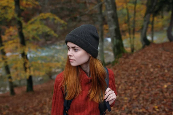 Femme énergique touriste avec un sac à dos dans un pull rouge et casquettes se reposent dans la forêt d'automne dans le parc — Photo