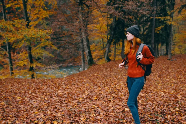 Kobieta w kapeluszu w czerwonym swetrze i dżinsach spaceruje po parku z plecakiem na plecach turystyka turystyczna jesienny krajobraz — Zdjęcie stockowe