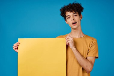 Kıvırcık saçlı, neşeli bir adam. Sarı posterli, mavi arka planlı.