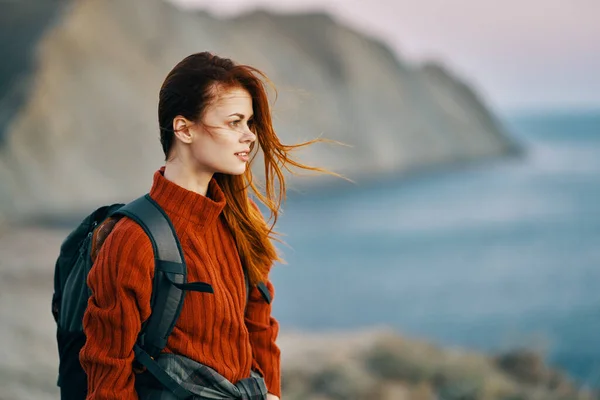 Viaggiatore dai capelli rossi con uno zaino sulla schiena in un maglione sulla spiaggia in montagna vicino al mare — Foto Stock