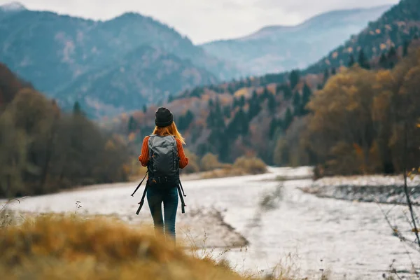 Viaggiatrice donna con zaino e cappello vicino al fiume in montagna in autunno — Foto Stock