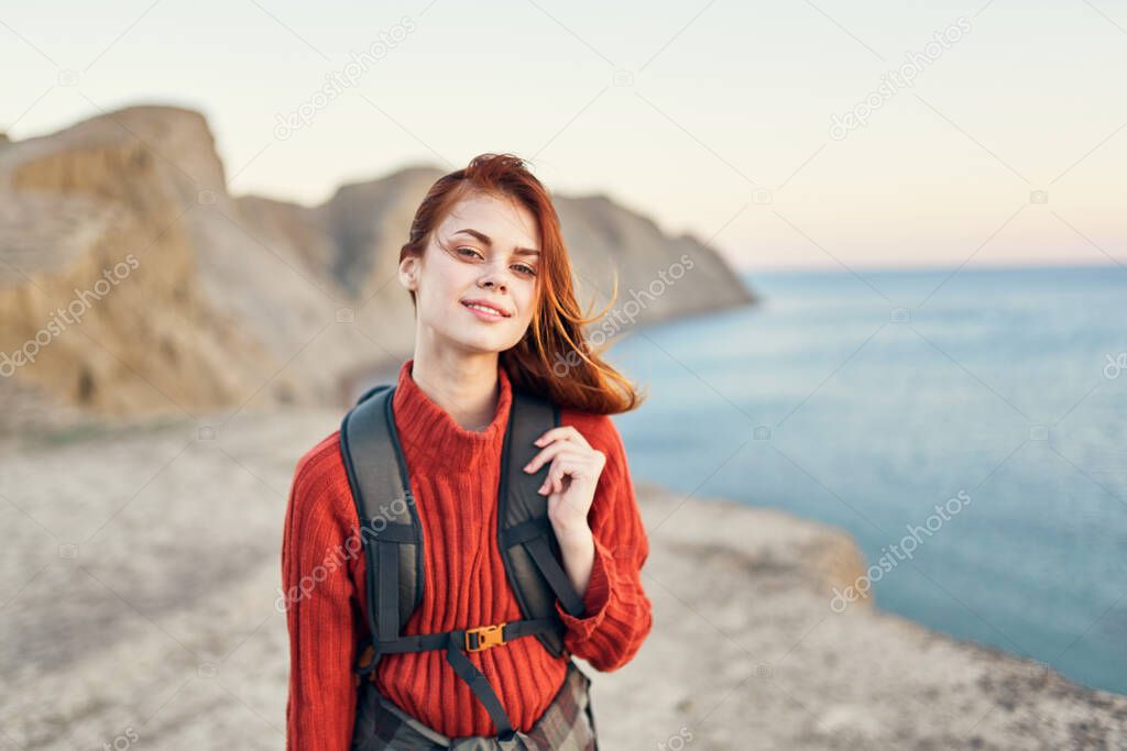 woman in a sweater with a backpack on her back near the sea in the mountains smile fun model