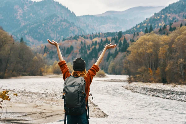 Een reiziger in een sweater hoed met een rugzak gebaren met haar handen en ontspant op de oever van de rivier in de bergen — Stockfoto
