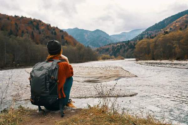 Femme dans les montagnes en automne avec un sac à dos Assis sur la rive de la rivière et regarde les hautes montagnes — Photo