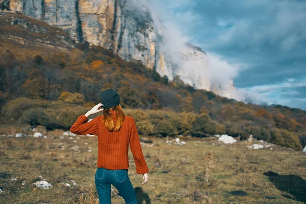 Una mujer en un suéter sostiene sus manos detrás de su cabeza y mira las altas montañas paisaje cielo azul nubes — Foto de Stock