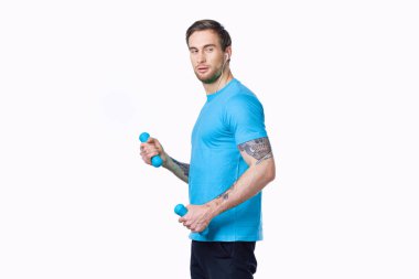 Mavi tişörtlü ve pantolonlu hafif arka planda dambıllarla egzersiz yapan bir adam.