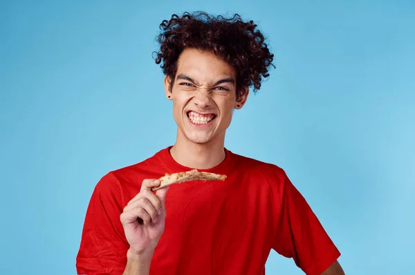 Energisk kille med en bit pizza har kul på en blå bakgrund och en röd t-shirt — Stockfoto