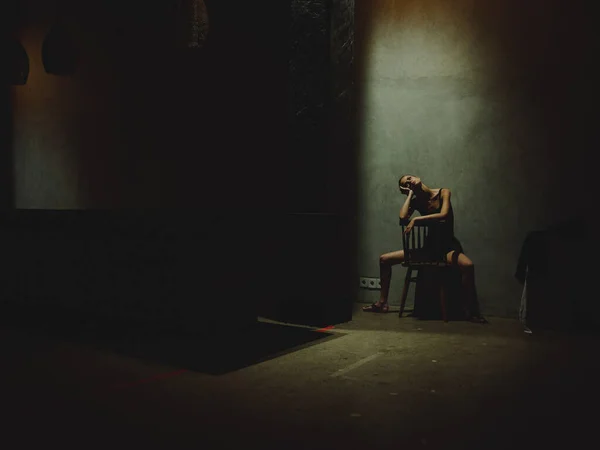 다리를 벌리고 어두운 방에 있는 의자에 앉아 위에서 불빛을 바라보는 한 여자 — 스톡 사진