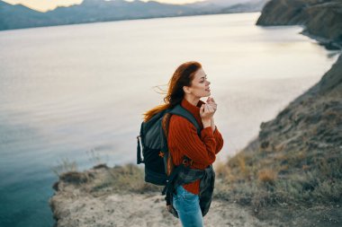 Deniz kenarındaki dağlarda sırt çantalı bir yürüyüşçü ve kırmızı bir kazak gevşek saçlı günbatımı kayaları.