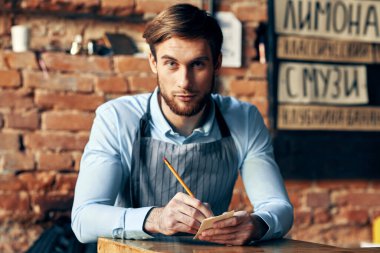 Önlüklü bir adam masada oturur ve bir garsonun siparişini kabul eder.