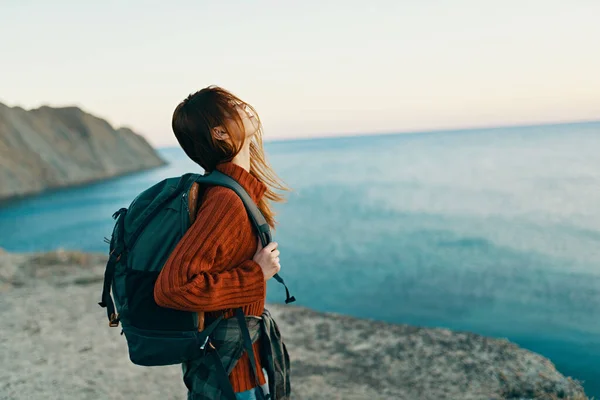 Viaggiatore felice in un maglione rosso con uno zaino sulla schiena guarda il mare in lontananza e in alta montagna — Foto Stock