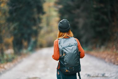 Sırt çantalı, şapkalı ve turuncu kazaklı bir kadın sonbahar ormanı manzaralı yolda.
