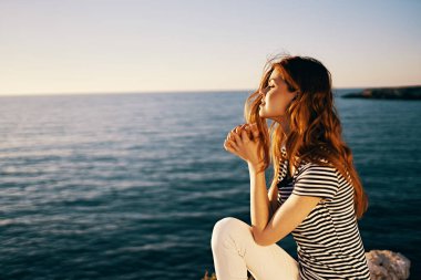 Çizgili tişörtlü ve beyaz pantolonlu bir kadın yazın deniz kenarında bir taşın üzerinde oturur.